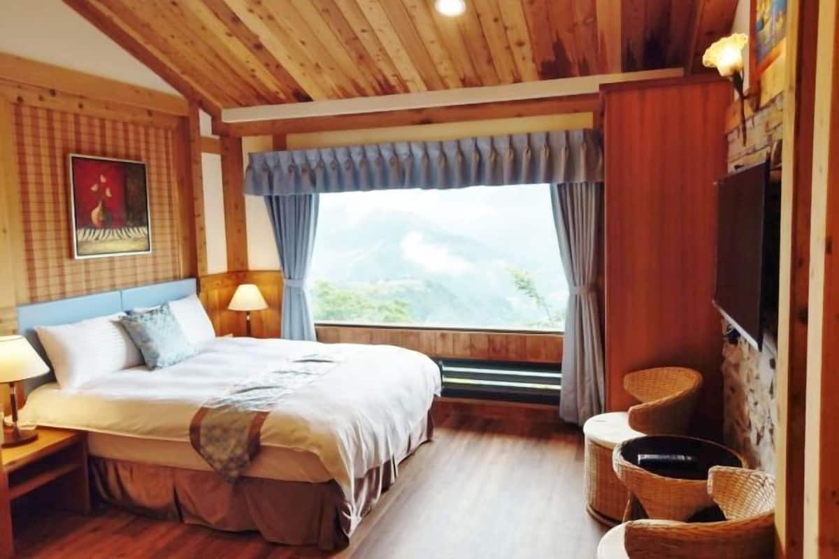A bedroom with a bed, bedside tables, a window with curtains, a wall-mounted TV, and wicker chairs.