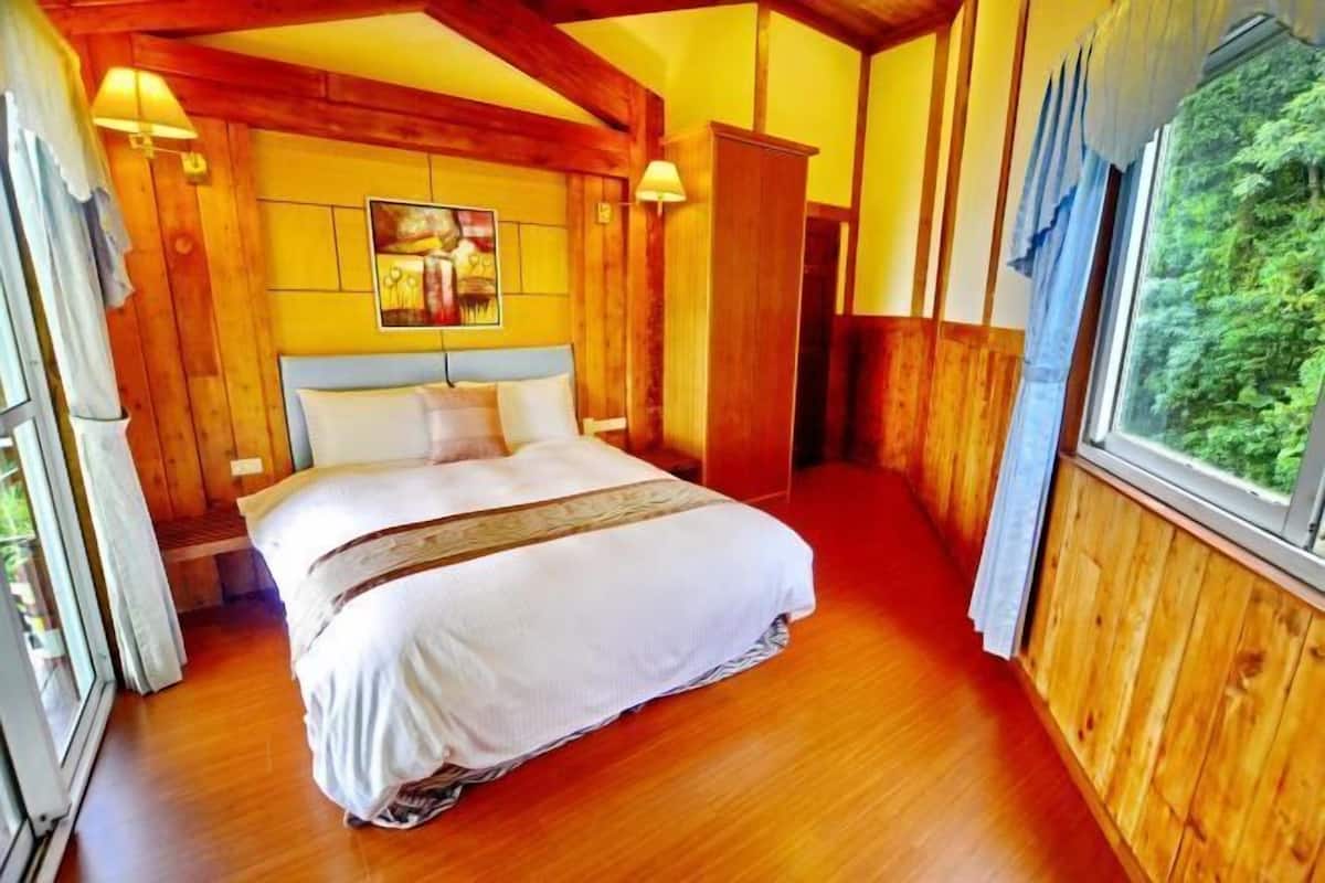 A wooden-paneled bedroom with a bed, a painting on the wall, and a window with a view of greenery.