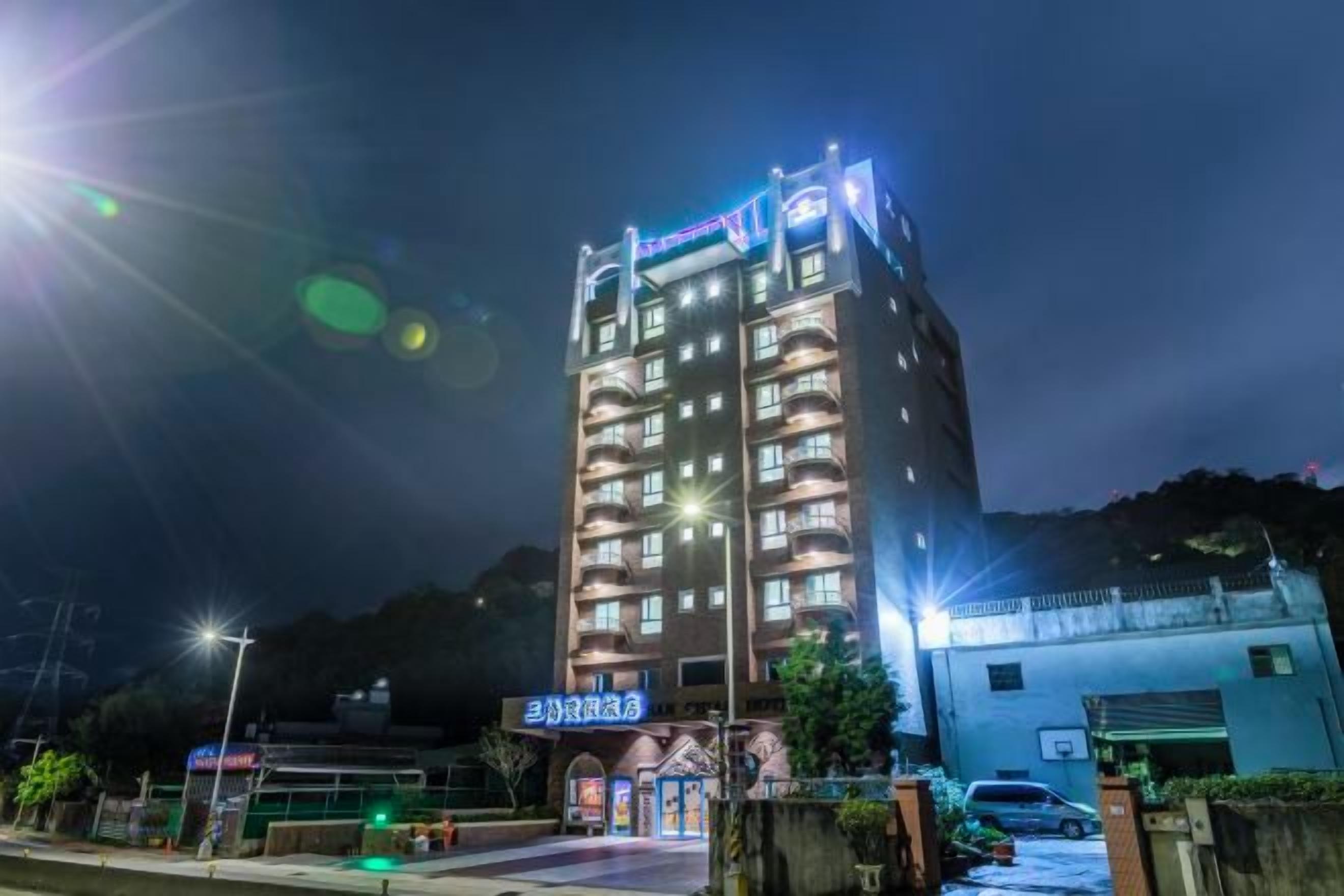 Hotel com vários andares, fachada iluminada, área de estacionamento e entrada com portão.