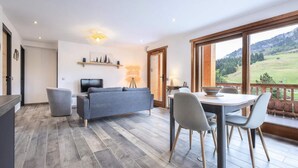 A modern living room with a grey sofa, a dining table with chairs, a TV, and a balcony with a scenic view. - 3 Rooms for 4 People (Le Grand-Bornand)