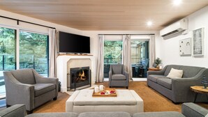 Een moderne woonkamer met grijze banken, een open haard, een flatscreen-tv en grote ramen met uitzicht op het omliggende landschap.