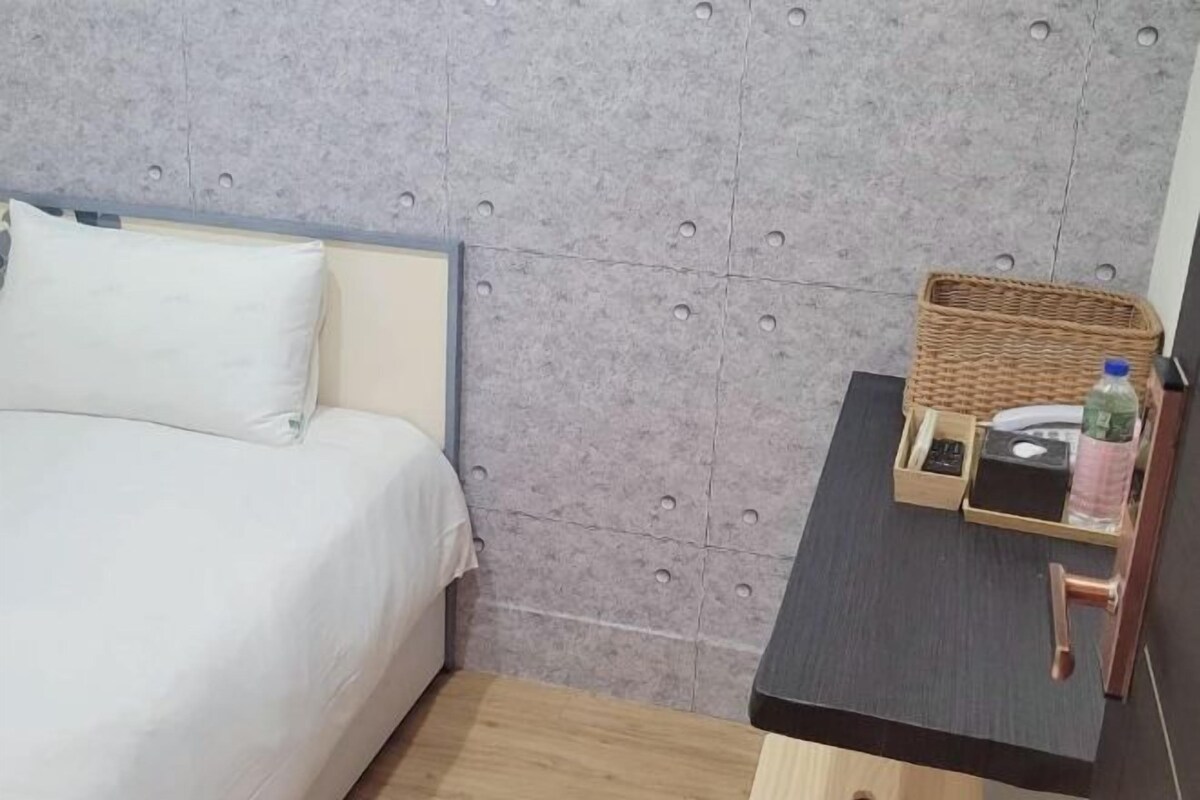 A bed with a headboard, a small table with a basket, a bottle, and a remote control.