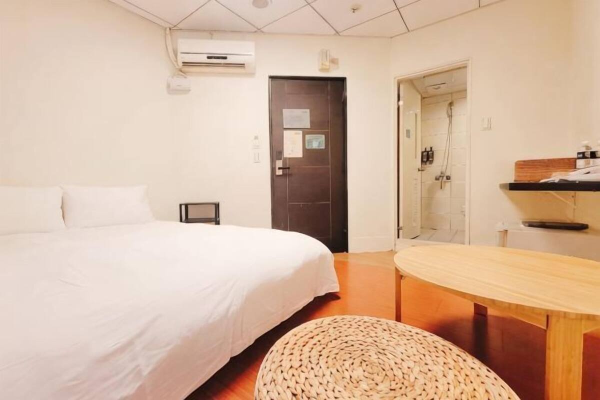 A hotel room with a bed, a wooden table, a woven stool, a door, and a shower area.