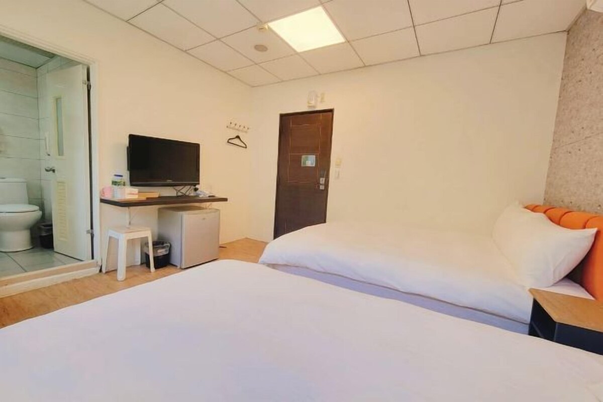 A hotel room with a bed, a desk with a television, a small stool, and a door.