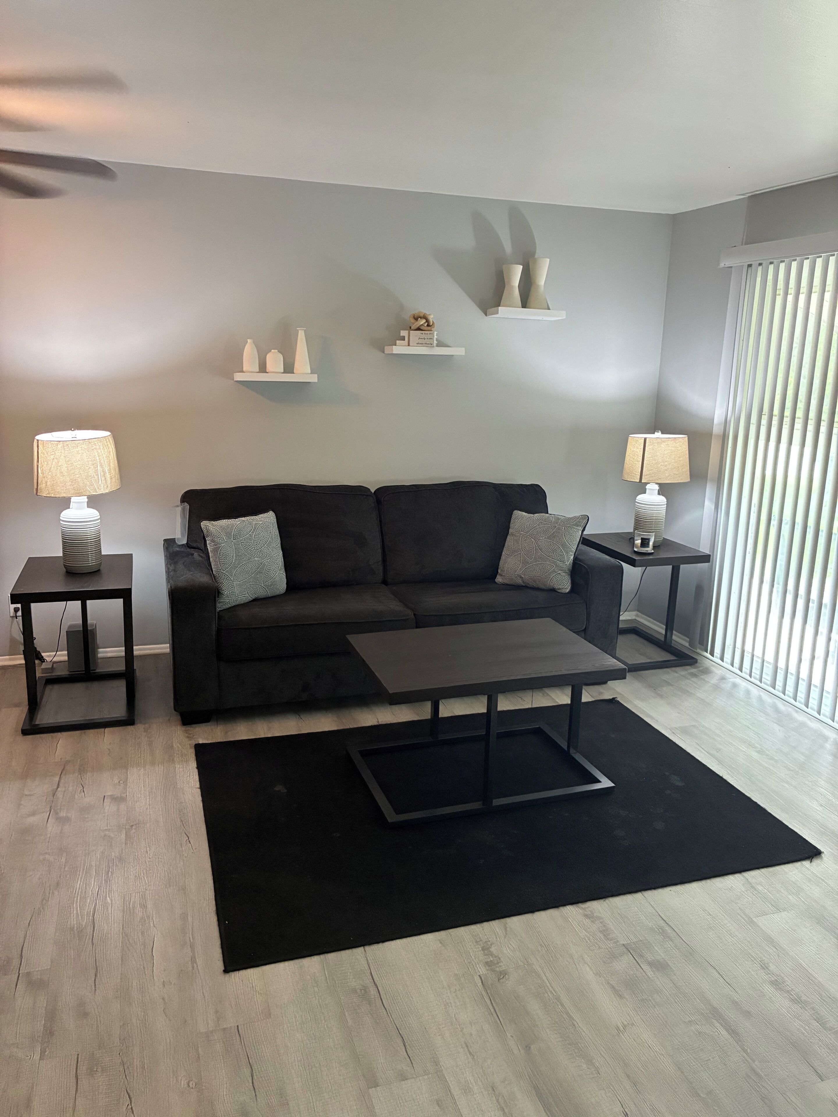 A modern living room with a black sofa, two side tables with lamps, a coffee table, and decorative shelves.
