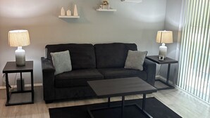 Un salon moderne avec un canapé noir, deux tables d’appoint avec des lampes, une table basse et des étagères décoratives.