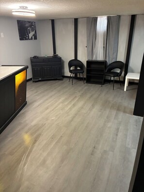 Una habitación con piso de madera, un gabinete negro, dos sillas negras, una mesa blanca y una guitarra negra en la pared.