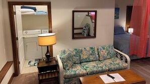 A hotel room with a sofa, a table with a remote control, a mirror, a bed with a red curtain, and a laundry area with a washing machine and dryer. - Beach Side HideAway - Pet Friendly, WiFi, Smart TV, Central Heat/AC, Parking (Norfolk)