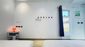 Habitación de hotel con dos camas, una mesita de noche con una lámpara roja, una ventana con cortinas azules y un perchero en la pared.