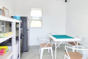 Une petite pièce avec une table et des chaises blanches, un réfrigérateur et une fenêtre.
