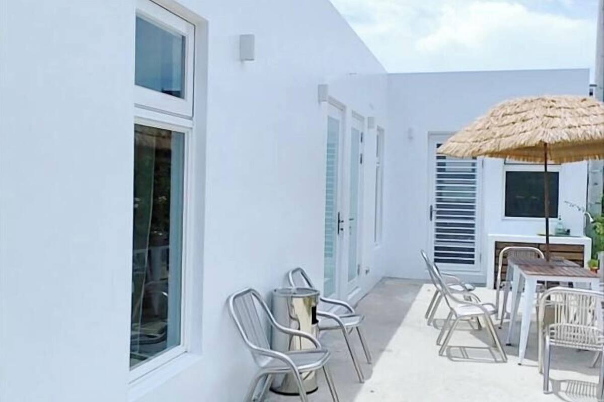 A white building with a patio area featuring a table and chairs, a thatched umbrella, and a trash can.