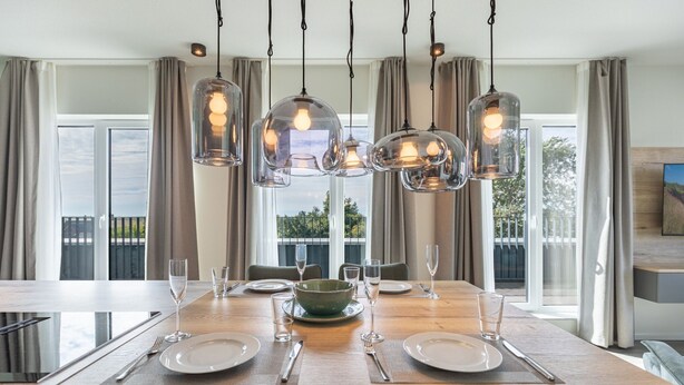 Un coin repas avec une table en bois dressée pour quatre personnes, entourée de chaises. Au-dessus, on trouve trois suspensions avec des ampoules apparentes. La pièce est dotée de grandes fenêtres avec des rideaux, et offre une vue sur la verdure à l'extérieur.