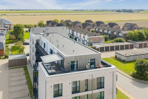 A modern multi-story building with a flat roof, large windows, and a flat roof terrace with outdoor seating. - Luxusapartement mit Meerblick (Nordstrand)