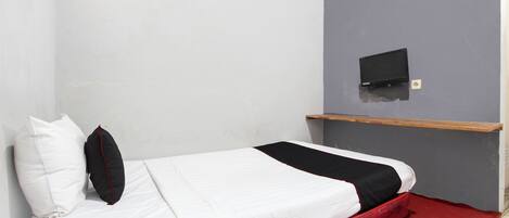 A single bed with a white and black bedspread, a red blanket, and two pillows (one white, one black) in a room with a grey wall and a small shelf with a black object on it.