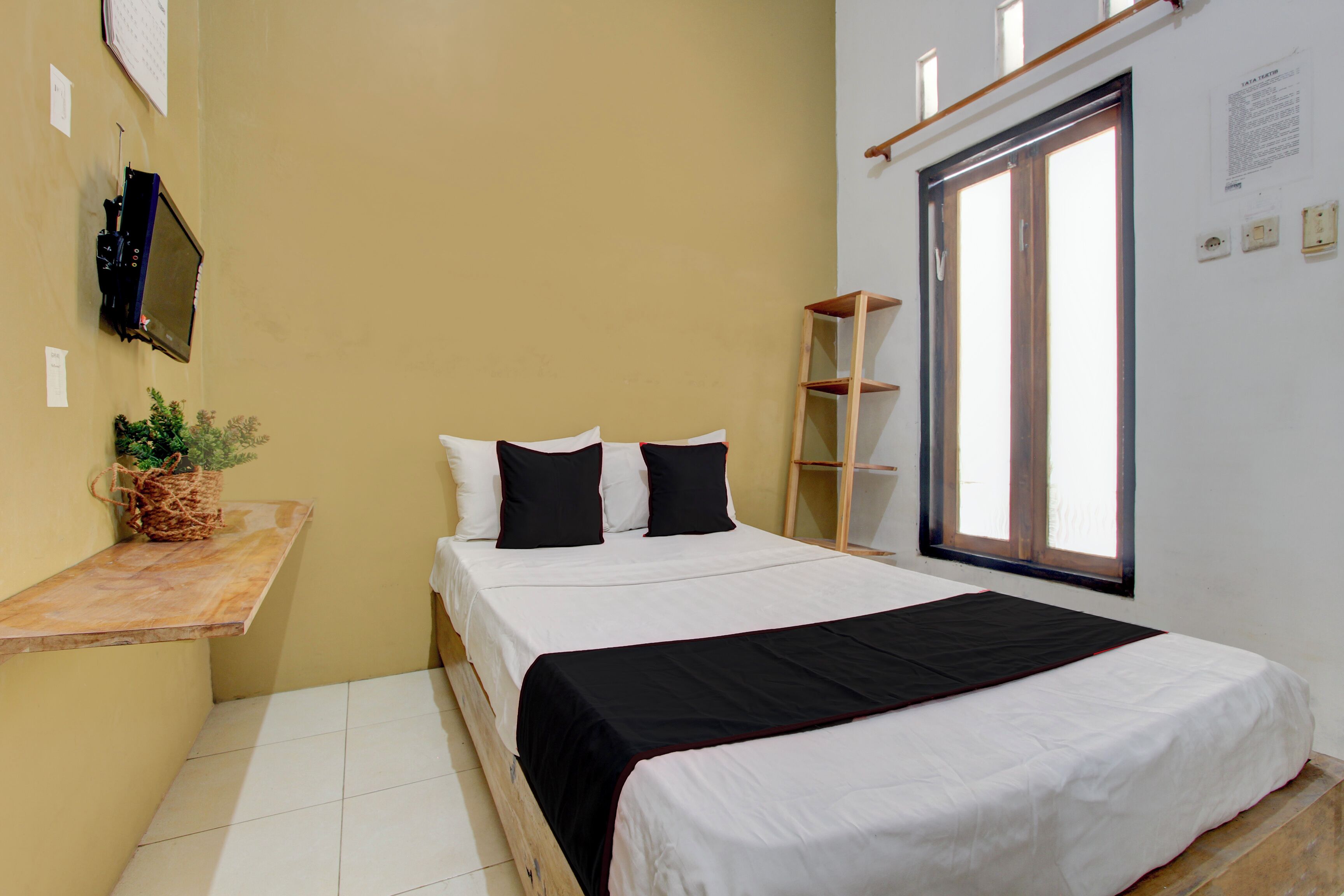 A bedroom with a bed, a shelf, a window, and a wall-mounted TV.