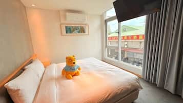 Cama com um urso de pelúcia, janela com vista para edifícios e televisão na parede.