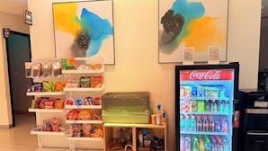 Un distributeur automatique proposant des en-cas et du Coca-Cola, une étagère remplie de divers articles et une œuvre d’art abstrait accrochée au mur.