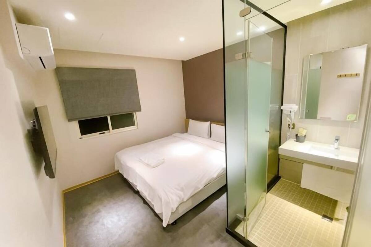 Une chambre à coucher comprenant un lit, une table de chevet, une fenêtre, un miroir, ainsi qu’une salle de bain avec un lavabo et un miroir.