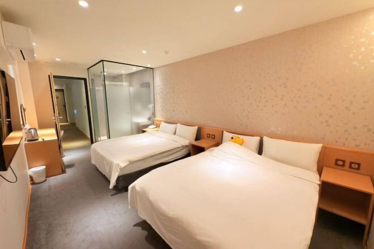 Une chambre d’hôtel avec deux lits, une cloison en verre, une table de chevet en bois et un petit bureau.