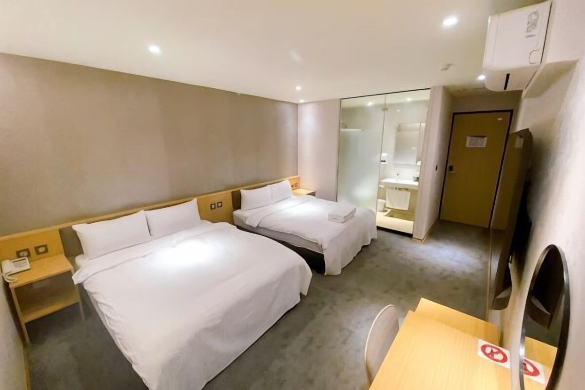 A hotel room with two beds, a desk, a chair, a mirror, a door, and a small table with a red sign.