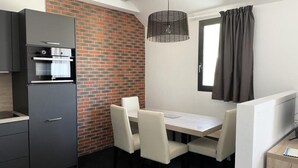 Een compacte hotelkamer met een kitchenette, een eettafel met stoelen, een raam met gordijnen en een accentmuur van baksteen.