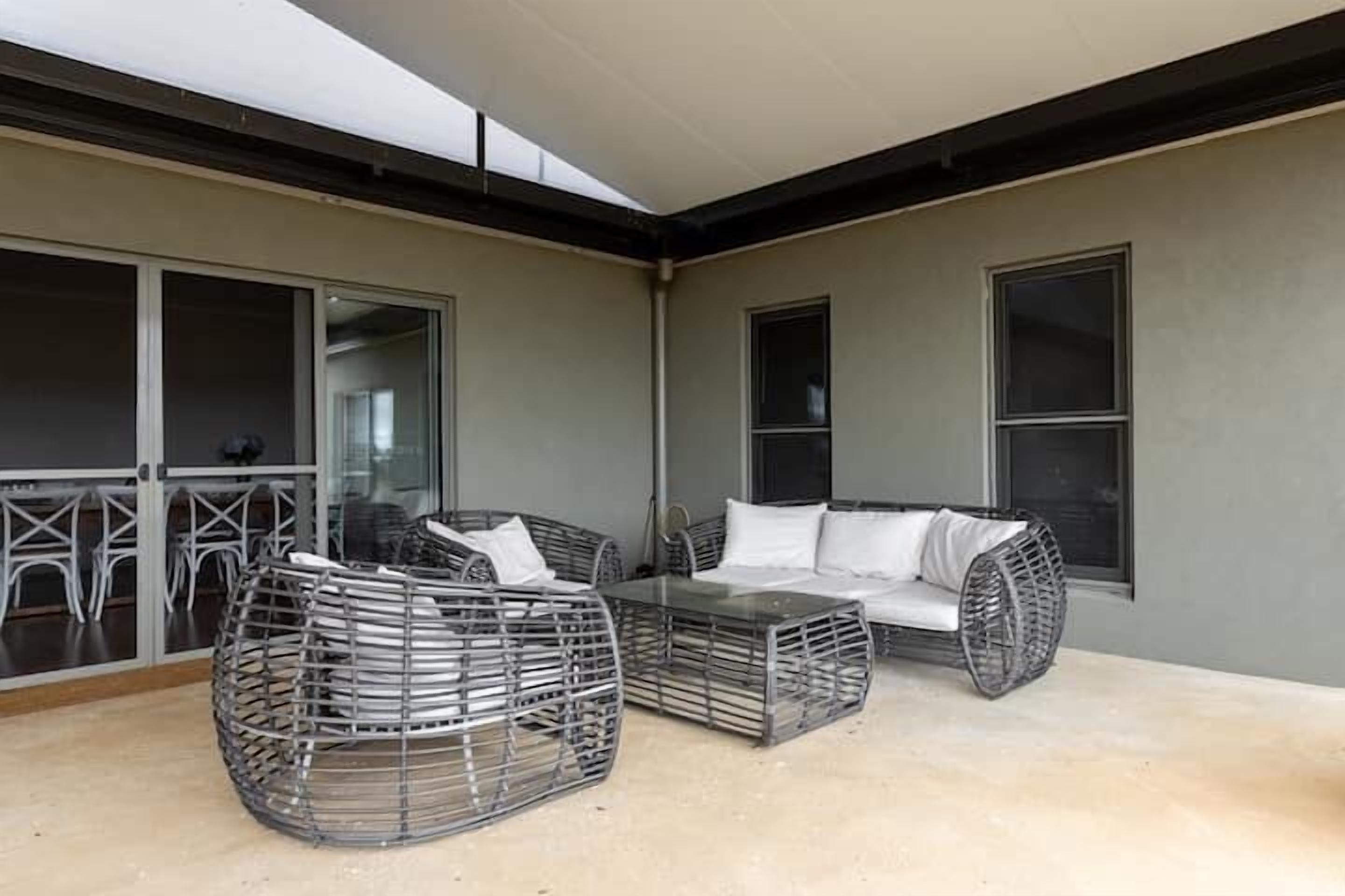 Een overdekt terras met rieten meubels en een glazen tafel, grenzend aan een schuifpui die uitkomt op een balkon.
