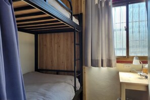 Una cama individual en literas con cabecero de madera, un escritorio con lámpara y una ventana con cortinas.
