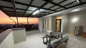 Eine Dachterrasse mit Tisch und Bänken, Blick auf die Stadt im Abendrot, und ein überdachter Bereich mit einer Leiter.