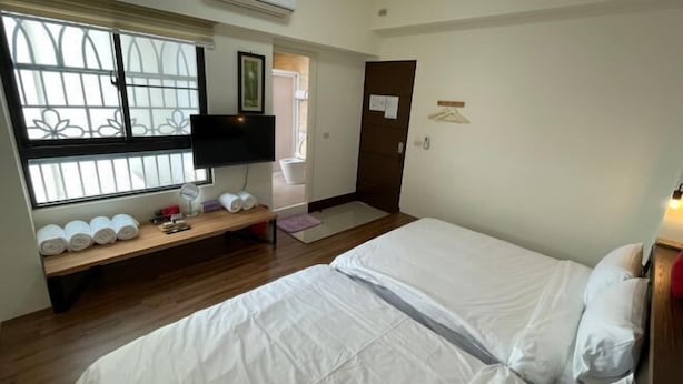Ein Hotelzimmer mit Bett, Fernseher, Fenster mit Jalousien, Nachttisch mit Handtüchern und einer Tür.