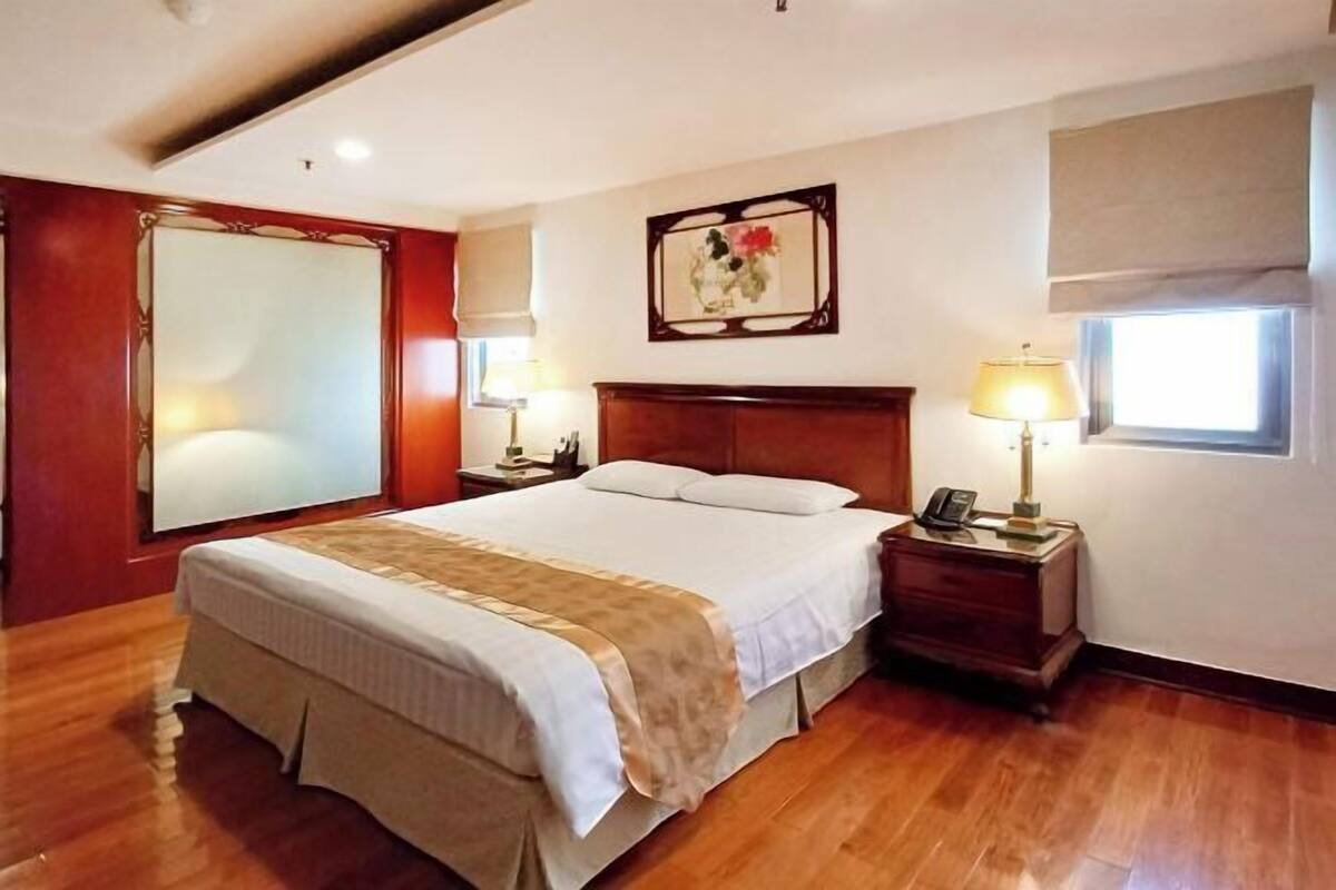 A hotel room with a large bed, wooden flooring, a mirror, bedside lamps, a nightstand, and a window with a view.