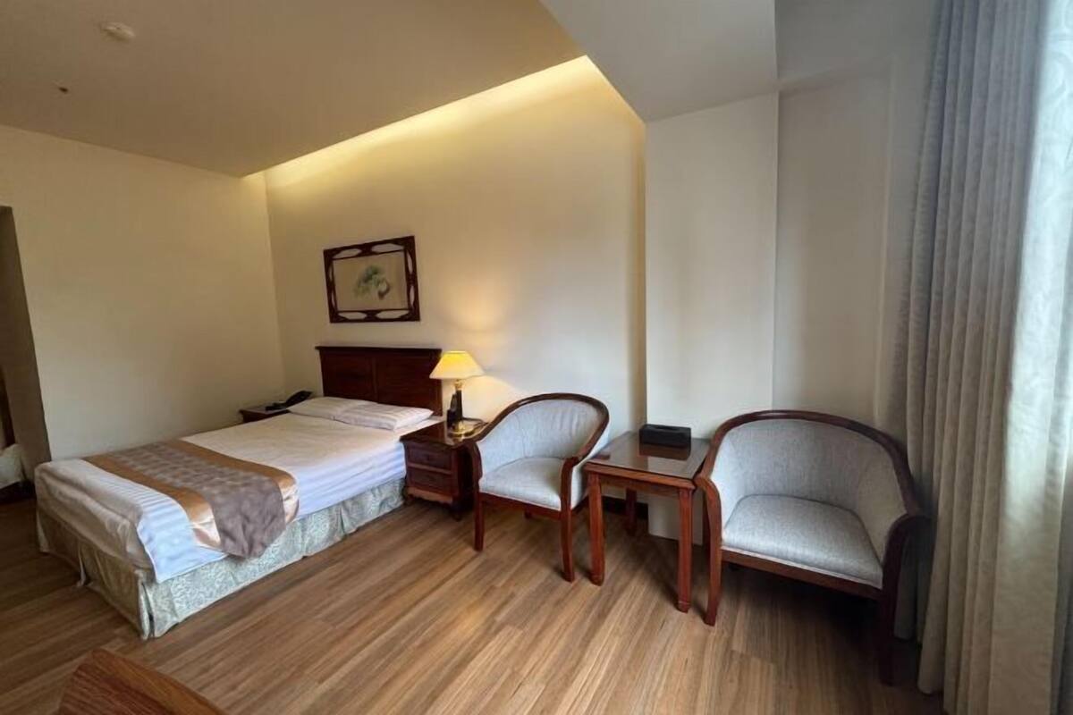 A hotel room with a bed, two chairs, a small table, a lamp, and a framed picture on the wall.