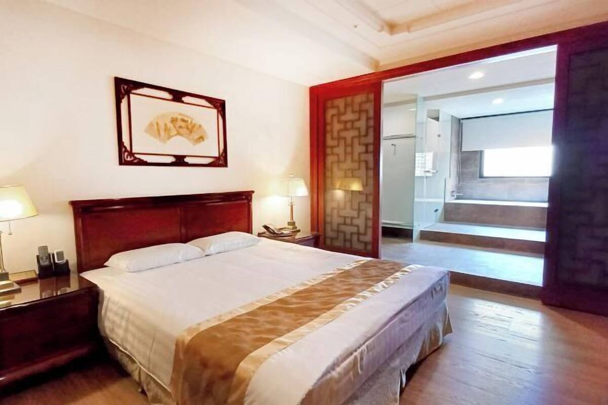 A hotel room with a bed, bedside tables, a wall-mounted picture, and a view of the kitchen through an open door.