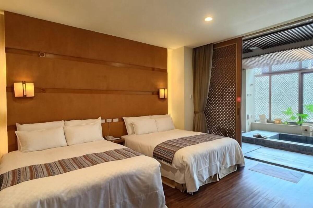 A hotel room with two beds, wooden headboard, wall-mounted lights, and a view of a garden through a sliding door.