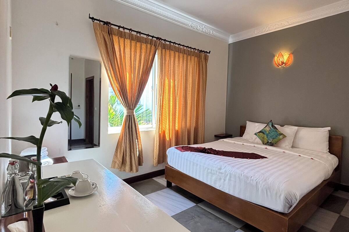 Una habitación de hotel con cama, escritorio, planta y ventana con cortinas.