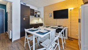 A compact kitchen with modern appliances, a dining area with white furniture, and a flat-screen TV mounted on the wooden wall. - Cozy Apartment Near the Slopes (Les Orres)