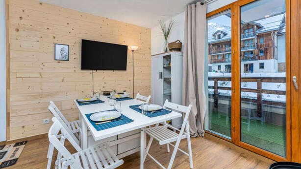 A dining area with a white table set for four, a television mounted on the wall, and a view of a snowy mountain through a glass door. - Cozy Apartment Near the Slopes (Les Orres)