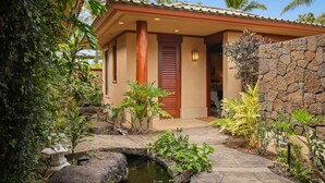 A house with a stone wall, a small pond, and tropical plants. - Maile Hale-Serenity Home (Waimea)