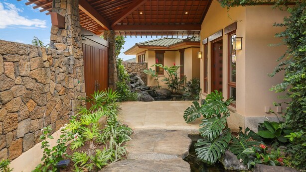 A covered stone pathway leading to a house with a stone wall on one side and a garden with tropical plants. - Maile Hale-Serenity Home (Waimea)