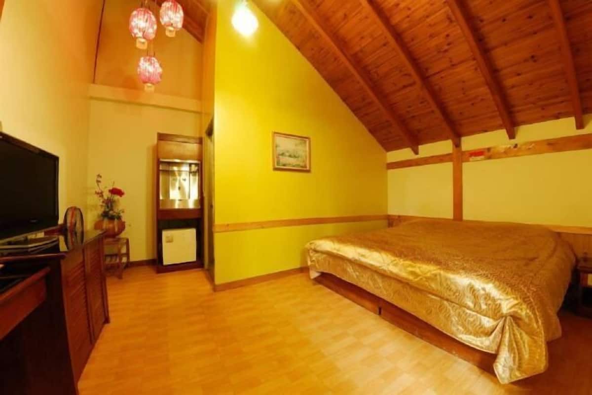 A bedroom with a wooden ceiling, a bed with a gold cover, a television, and a small table with a vase of flowers.