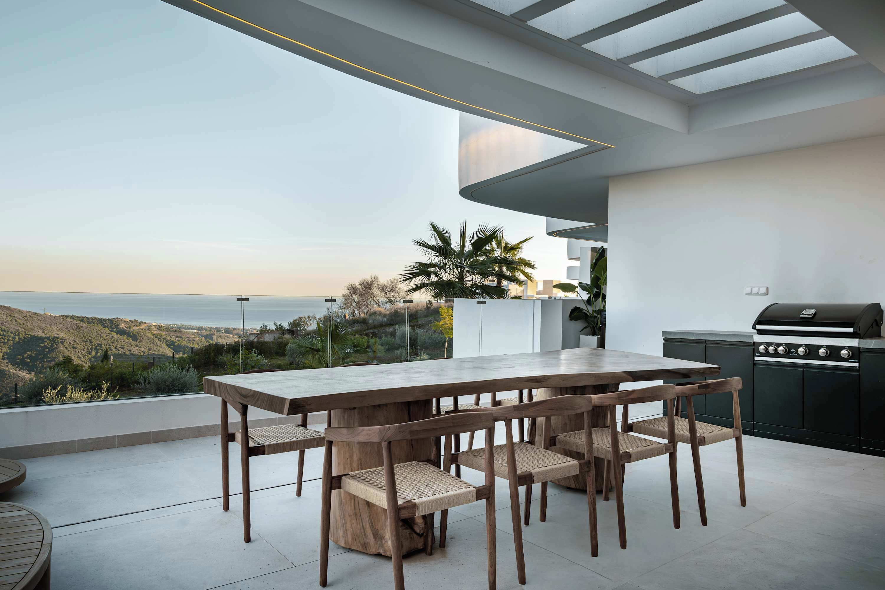 Un área de comedor exterior moderna con una mesa grande y sillas, una parrilla y vistas a las colinas.