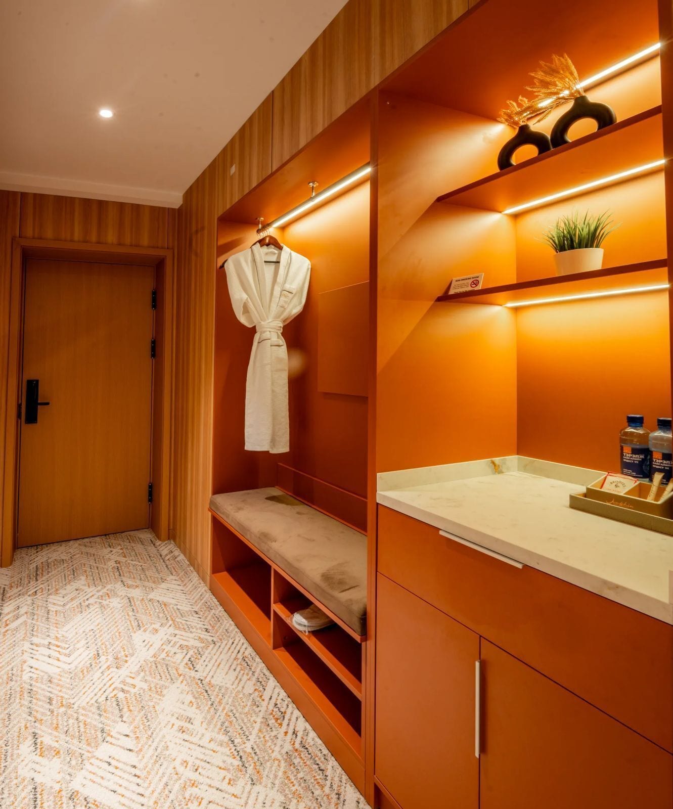 A hotel room with a wooden wardrobe, a bench, a robe, a sink, and a small plant.