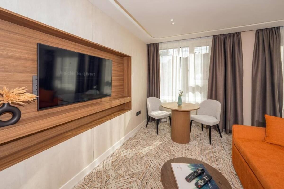 Una habitación de hotel moderna con una televisión de pantalla plana montada en un panel de madera, una mesa redonda con dos sillas, un sofá y una mesa baja con mandos a distancia.