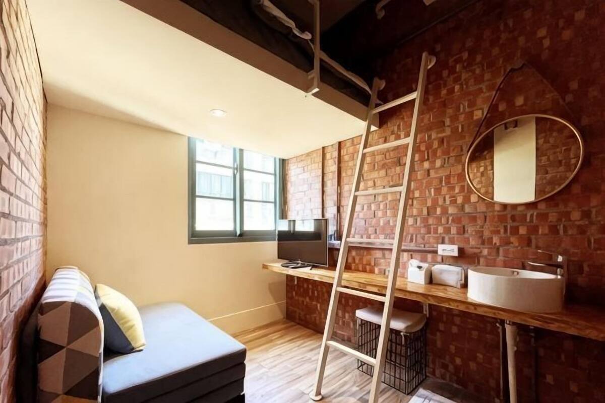 Une pièce de taille modeste avec un mur en briques, un plancher en bois, un canapé, une échelle, un évier et un miroir.