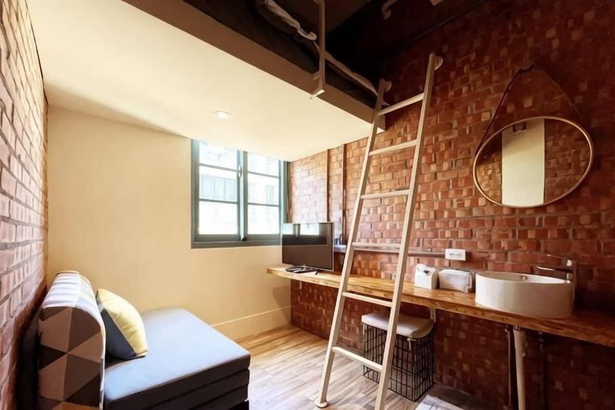 A compact room with a brick wall, a wooden floor, a sofa, a ladder, a sink, and a mirror.