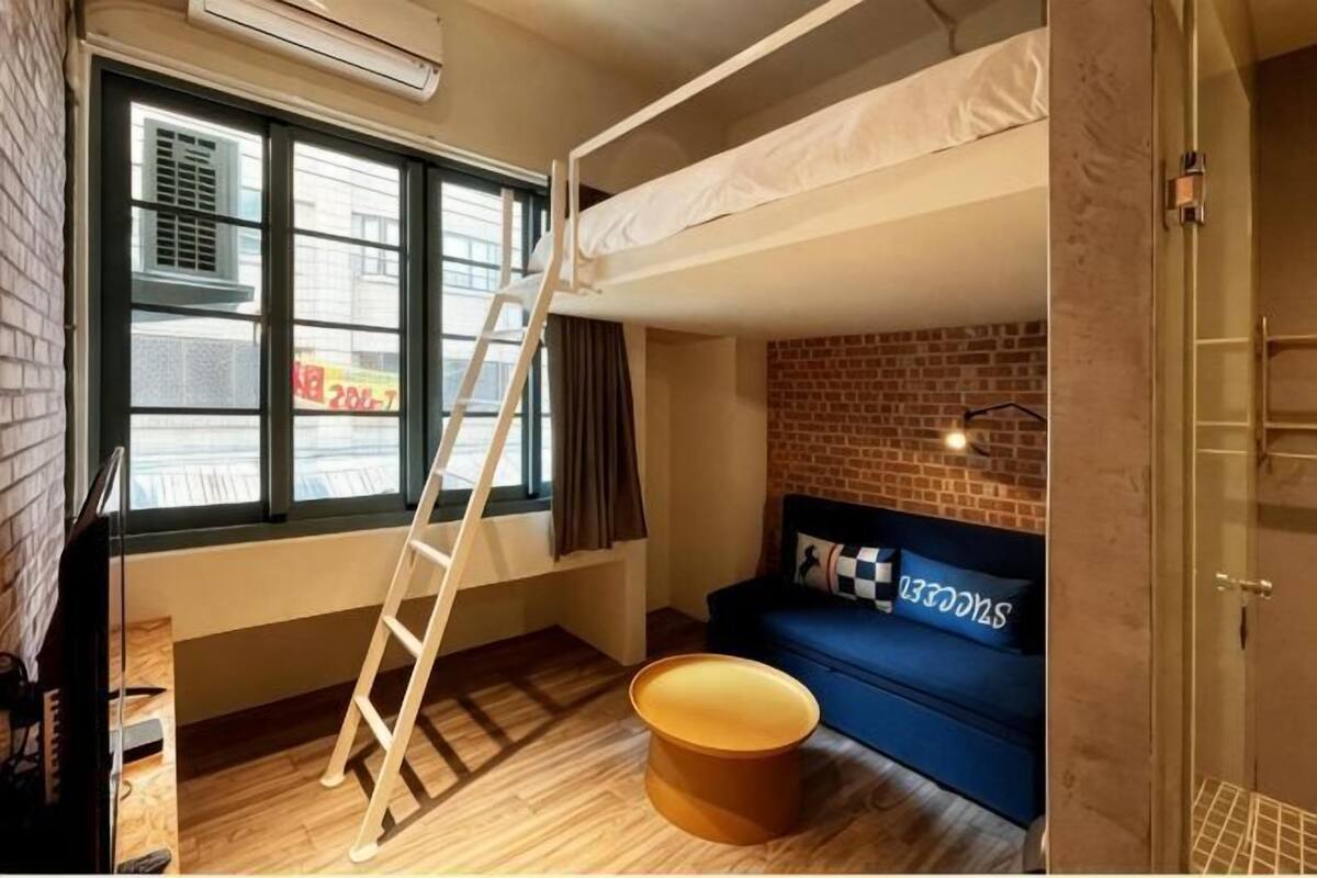 Une pièce avec un lit superposé, un canapé, un pouf rond, une échelle, une fenêtre et un mur en briques.