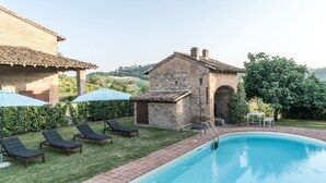 A swimming pool with lounge chairs and umbrellas, a brick building, and a view of the countryside.