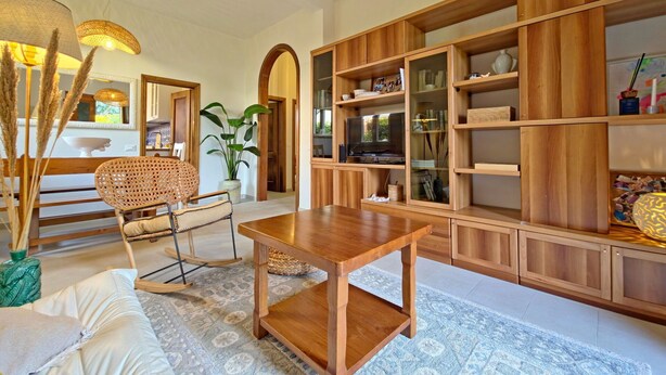 Un vaste espace de vie agrémenté d’une table basse en bois, d’un fauteuil en rotin et d’une grande bibliothèque remplie de livres et d’objets décoratifs.
