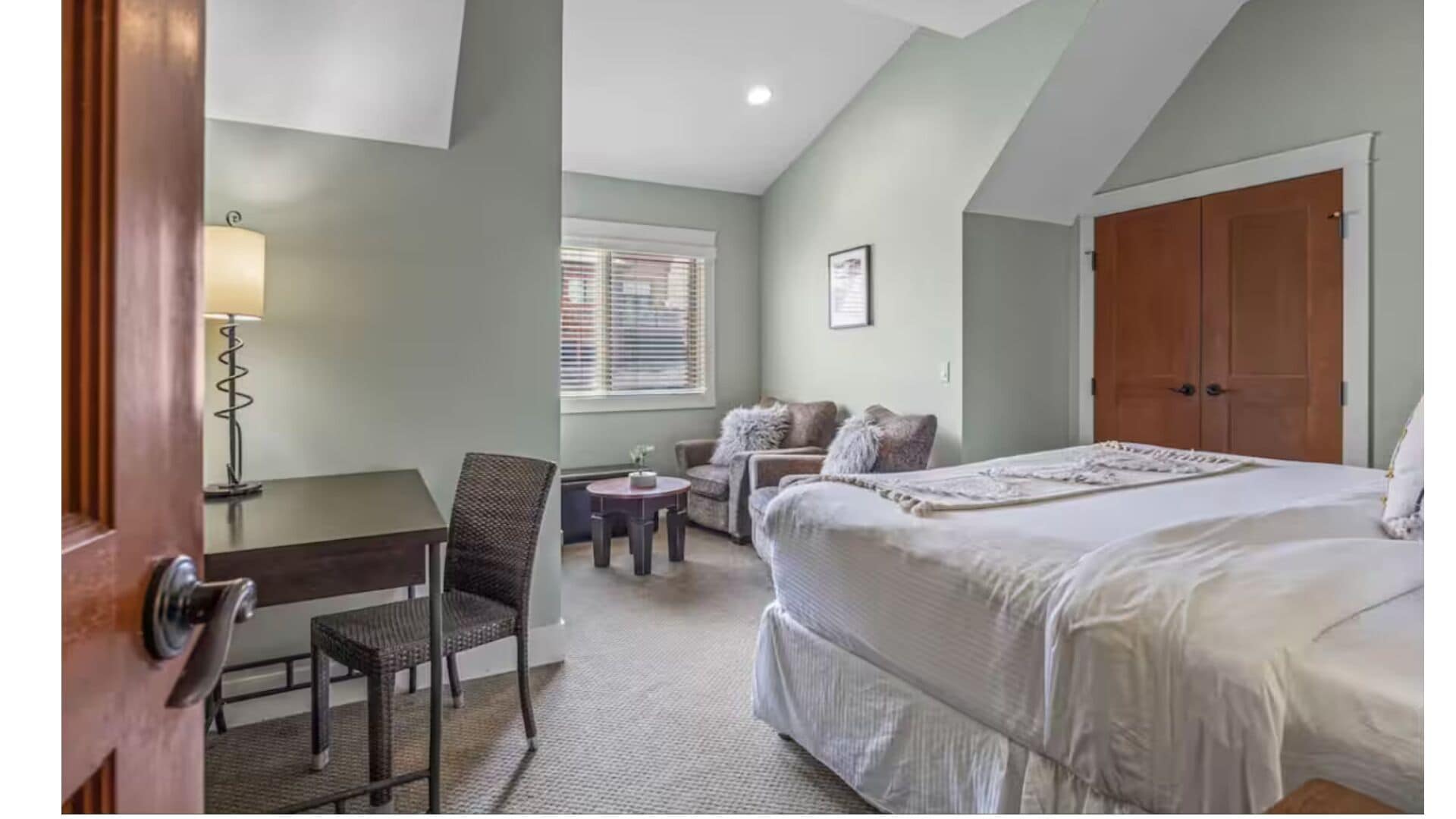 A bedroom with a large bed, a desk, a chair, and a window with blinds.