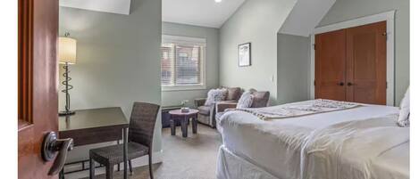 A bedroom with a large bed, a desk, a chair, and a window with blinds.