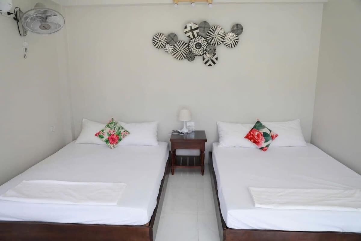 A hotel room with two beds, a small table, a wall-mounted fan, and a decorative wall piece.